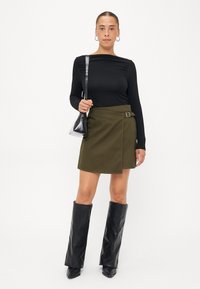 Zwarte lange mouwen top, olijfgroene wikkelrok met gespdetail, knie hoge zwarte lakleren laarzen, kleine zwarte handtas.
