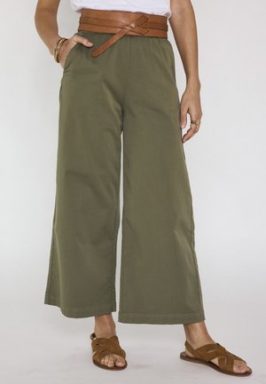 Persona che indossa pantaloni corti a gamba larga color oliva con cintura marrone a fascia e sandali di pelle marrone, in piedi con una mano in tasca.