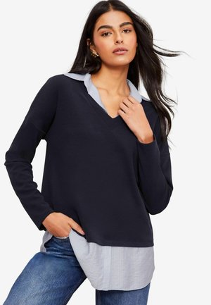 Marineblå v-hals top med lange ærmer over en lyseblå stribet skjorte med knapper. Blødt stof, afslappet pasform, asymmetrisk kant og casual stil.