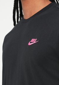 Juodas medvilnės marškinėliai trumpomis rankovėmis su rožiniu siuvinėtų Nike logotipu krūtinėje, standartiniu apvaliu kaklu. Lygus paviršius.