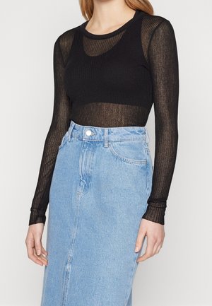 Femme portant un haut noir transparent à manches longues avec un débardeur noir sans manches, associé à une jupe en jean bleu clair taille haute.