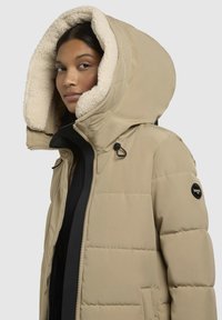 Bežs pūkains jaka ar lielu, mīksti izklātu kapuci, rāvējslēdzēju, sānu kabatām un melnu logo uzlīmi uz piedurknes.