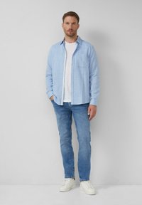 Hellblaue karierte Langarmbluse mit einer Brusttasche, über einem weißen Rundhals-T-Shirt. Trägt dazu hellblaue Jeans und weiße Sneaker.