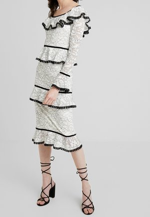 Femme portant une robe en dentelle blanche avec un bord noir et des détails pompons, associée à des sandales noires à talons hauts et à lanières.