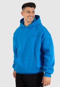 Uomo sorridente, che indossa una felpa sportiva Smilodox di un brillante blu con tasca anteriore e pantaloni neri, in piedi davanti a uno sfondo grigio chiaro.