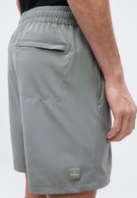 Άνδρας που φοράει γκρι σορτς με ελαστική μέση και πίσω τσέπη, με λογότυπο Calvin Klein στο πόδι, στέκεται με το δεξί χέρι χαλαρό στο πλάι.