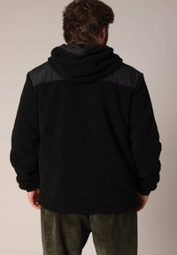 Zwarte fleecjas met een textuur oppervlak, capuchon en contrasterende zwarte schouderpanelen. Heeft een drop shoulder-stijl en lange mouwen.