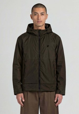 Uomo che indossa una giacca con cappuccio verde scuro con zip e pantaloni marroni, in piedi davanti a uno sfondo chiaro e uniforme.