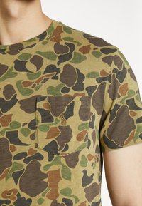 Tričko s krátkým rukávem v maskáčovém vzoru obsahující zelené, hnědé a černé odstíny. Obsahuje náprsní kapsu na levé straně s jemným prošíváním.