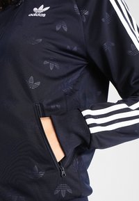Navy Adidas jacka i träningsstil med en texturerad design och subtila logotypmönster, en dragkedjeficka och vita tre ränder på ärmarna.