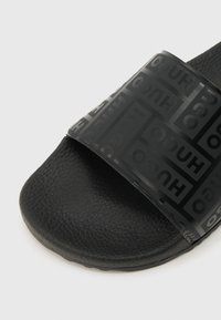 Κοντινό πλάνο μαύρου σανδαλιού τύπου σαγιονάρας με υφασμένη σόλα και φαρδιά λουράκι που φέρει επαναλαμβανόμενο λογότυπο "HUGO" σε πιο σκούρο μαύρο.