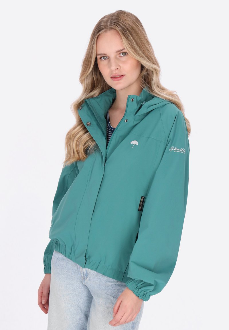 Lekka kurtka w kolorze morskim z kapturem, elastycznymi mankietami i regulowanym dołem. Posiada małe logo oraz przednią kieszeń na zamek.