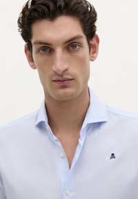 Lichtblauwe button-down shirt gemaakt van stof met een gladde textuur, met een klein geborduurd schedel logo op de linkerborst.