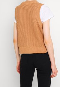 Gebreide mouwloze trui in lichtbruin met een gestructureerd patroon, geribbelde halslijn en zoom, gecombineerd met strakke zwarte jeans.