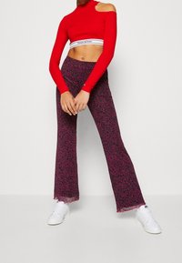 Punainen pitkähihainen lyhyt toppi, jossa on leikattu olkainten yksityiskohta, yhdistettynä tummanliilaisiin leveälahkeisiin housuihin, joissa on vaaleanpunainen abstrakti kuvio ja repaleiset helmat.