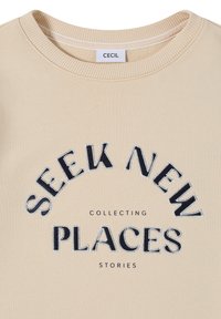 Sweat-shirt beige à col rond avec le texte "SEEK NEW PLACES" en grandes lettres brodées et un texte plus petit "COLLECTING STORIES" sous l'étiquette de la marque "CECIL".