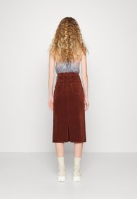 Falda midi de pana marrón con una abertura trasera, con cintura alta y dos bolsillos traseros, combinada con una camiseta gris claro y zapatillas beige sin cordones.