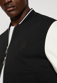 Chaqueta bomber negra con mangas blancas, con cierre de cremallera y un sutil logo de tigre en relieve en el pecho. Tejido suave y cuello acanalado.