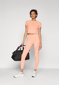 Pfirsichfarbener Crop-Top mit kurzen Ärmeln und Knopfdetails, kombiniert mit passenden hochgeschnittenen Leggings mit Ausschnitten, ergänzt durch eine schwarze Sporttasche.