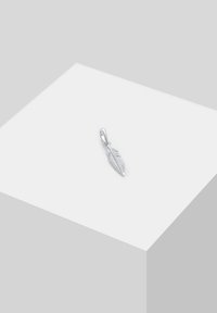 Petit pendentif en forme de plume argentée avec des lignes gravées détaillées, posé sur un socle cubique blanc sur fond gris.