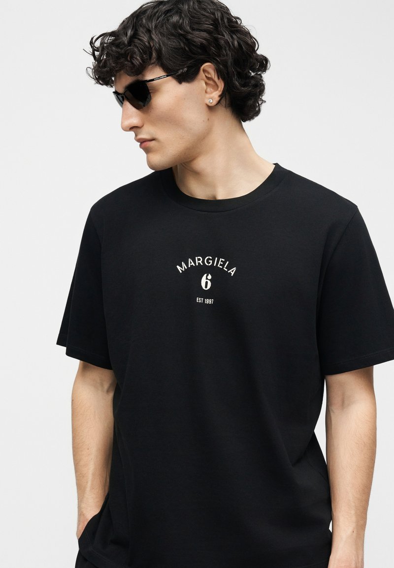 Mann mit lockigem, dunklem Haar und Sonnenbrille, trägt ein schwarzes T-Shirt mit der Aufschrift "MARGIELA 6 EST 1997" auf der Vorderseite, Hände in den Taschen.