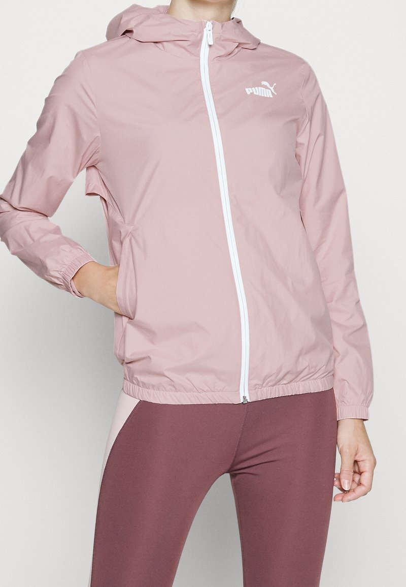 Femme portant une veste à capuche Puma rose clair avec une fermeture éclair blanche et des leggings bordeaux, main reposant dans la poche de la veste.