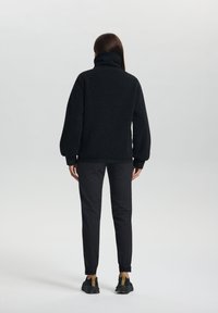 Zwarte fleece pullover met hoge kraag, verlaagde schouders en elastische manchetten, gecombineerd met slim-fit zwarte broeken en zwarte sneakers.