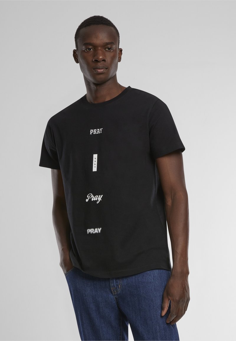 Young man wearing black t-shirt with multiple "PRAY" text prints in different fonts, paired with blue jeans, standing against gray background.