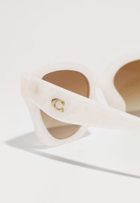 Üks suurte valgete plastist päikeseprillid geomeetrilise mustriga, pruuni läätsega ja kuldse logo rõhutusega äärisel.