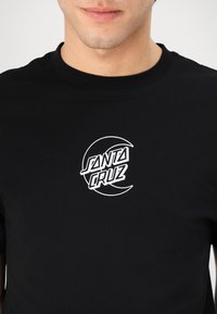 Juodas medvilnės marškinėlis su balta logo, kurioje užrašyta "SANTA CRUZ" menišku šriftu, sutelktas ant krūtinės. Trumpomis rankovėmis ir apvaliu kaklu.