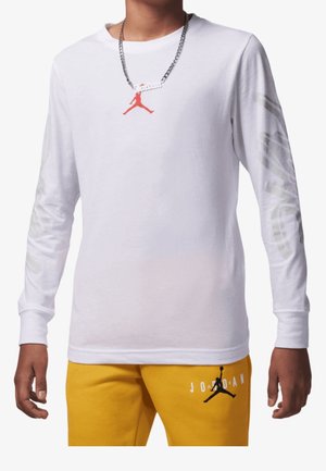 Personne portant un t-shirt Jordan blanc à manches longues avec logo rouge sur la poitrine et pantalon Jordan jaune avec logo noir sur la cuisse.