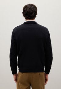 Uomo di spalle che indossa un maglione di maglia nera con maniche a motivo e pantaloni marroni, su uno sfondo chiaro e uniforme.