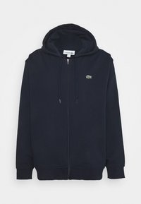 Felpa con zip di colore blu navy realizzata in tessuto misto cotone, dotata di tasca a marsupio, cappuccio con cordino e un piccolo logo verde sul petto.