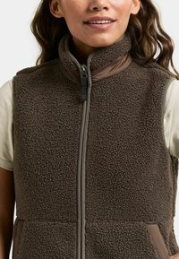 Vrouw draagt een bruin, gestructureerd fleecevest met een ritssluiting aan de voorkant over een lichtbeige T-shirt met korte mouwen, glimlacht lichtjes met zichtbare lippen en tanden.