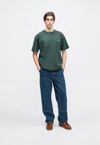 Tmavě zelené bavlněné tričko s kulatým výstřihem, doplněné volnými modrými denimovými džíny. Hnědé šněrovací boty outfit dotvářejí.