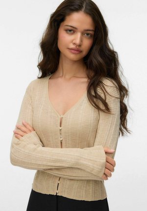 Młoda kobieta z długimi ciemnymi włosami, ubrana w beżowy prążkowany kardigan z perłowymi guzikami, z założonymi rękami, o neutralnym wyrazie twarzy na białym tle.