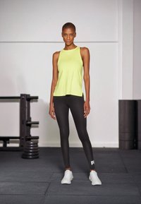 Débardeur de sport sans manches vert lime et leggings noirs, associés à des chaussures de sport blanches ; design épuré avec détail du logo sur les leggings.