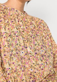 Blusa floreale in tessuto giallo con fiori rosa, viola e bianchi. Presenta un colletto rotondo e piccoli bottoni dorati lungo la parte frontale.