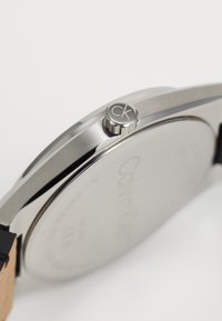 Reloj de acero inoxidable con correa de cuero negro. Presenta una corona plateada texturizada marcada con "CK" y un fondo de caja suave y reflectante.