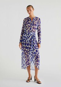 Abito lungo trasparente a maniche lunghe in stampa leopardata blu e viola, con scollatura bottonata, cintura in vita e orlo asimmetrico. Sandali neri con tacco.