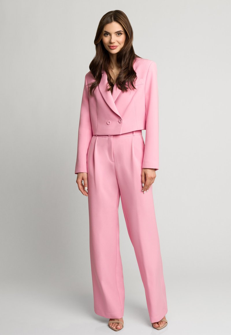 Różowy garnitur skrojony na miarę, z krótką marynarką dwurzędową i szerokimi spodniami. Gładki materiał, zorganizowany fason i subtelne fałdy.
