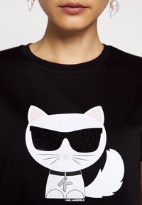 Camiseta negra de algodón con un diseño de un gato de dibujos animados en gafas de sol, con detalles de lentejuelas, mostrando una expresión juguetona y un collar con una llave.