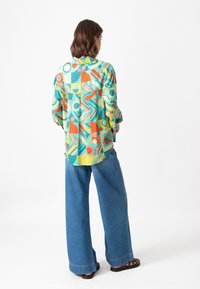 Camisa oversized de estampado colorido con formas geométricas en verde, naranja, amarillo y azul, combinada con pantalones de mezclilla de pierna ancha.