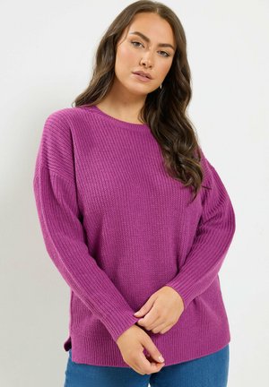 Kvinde med langt mørkt hår iført en struktureret lilla sweater og blå jeans, stående med hænderne blidt foldet foran sig.