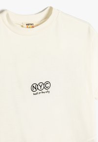 T-shirt de algodón crema con cuello redondo, mangas cortas y un diseño impreso con "NYC" y "mejor de la ciudad" en texto negro.