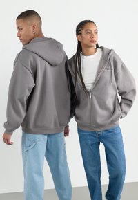 Grå jakker med glidelås, båret av to personer, med ribbestrikkede mansjetter, litt oversized passform, kombinert med lyseblå jeans.