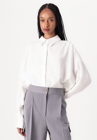 Chemise blanche oversize à col pointu ; associée à un pantalon gris clair à rayures avec un bouton à l'avant et des plis.