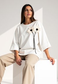 Biała, oversize'owa koszulka z bawełny z dwoma materiałowymi stokrotkami z czarnymi środkami i długimi czarnymi łodygami, zestawiona z beżowymi spodniami.