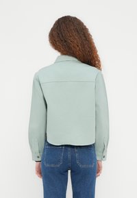 Veste courte vert menthe en tissu texturé, avec un col et des manches à boutons, associée à un jean bleu taille haute.