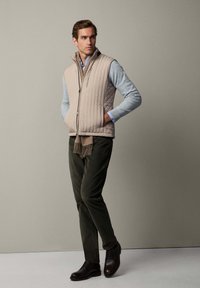 Gilet imbottito beige sopra un maglione blu chiaro, pantaloni verde scuro e scarpe marroni. Una sciarpa marrone con frange è indossata attorno al collo.